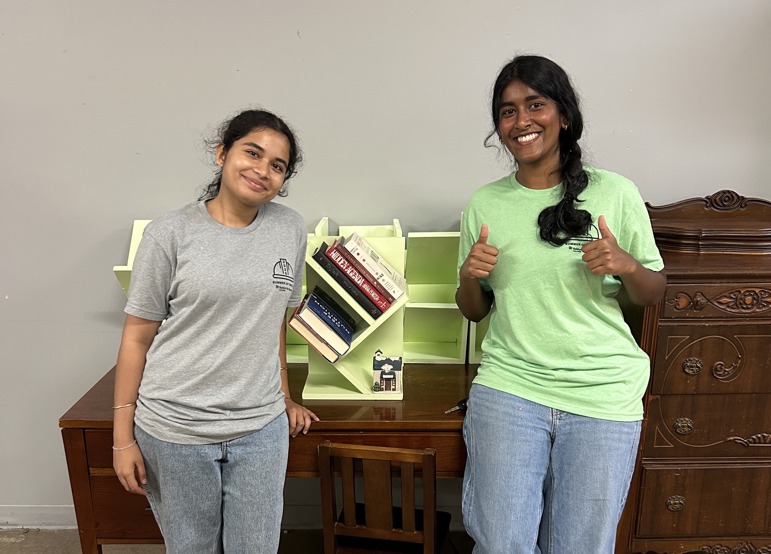 Two summer of service interns pose in front of bookshelf donations at the cary ReStore
